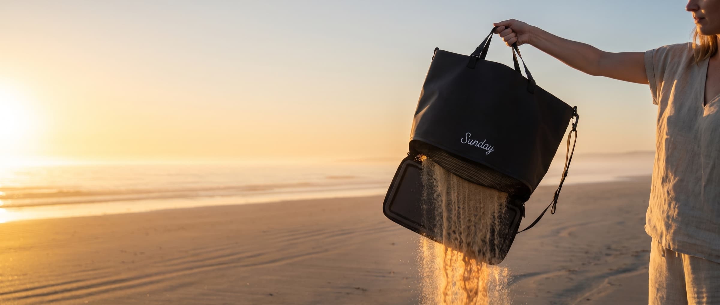 The Sunday Tote held over wet sand at golden hour, base open, backlit sand pouring out through the mesh floor